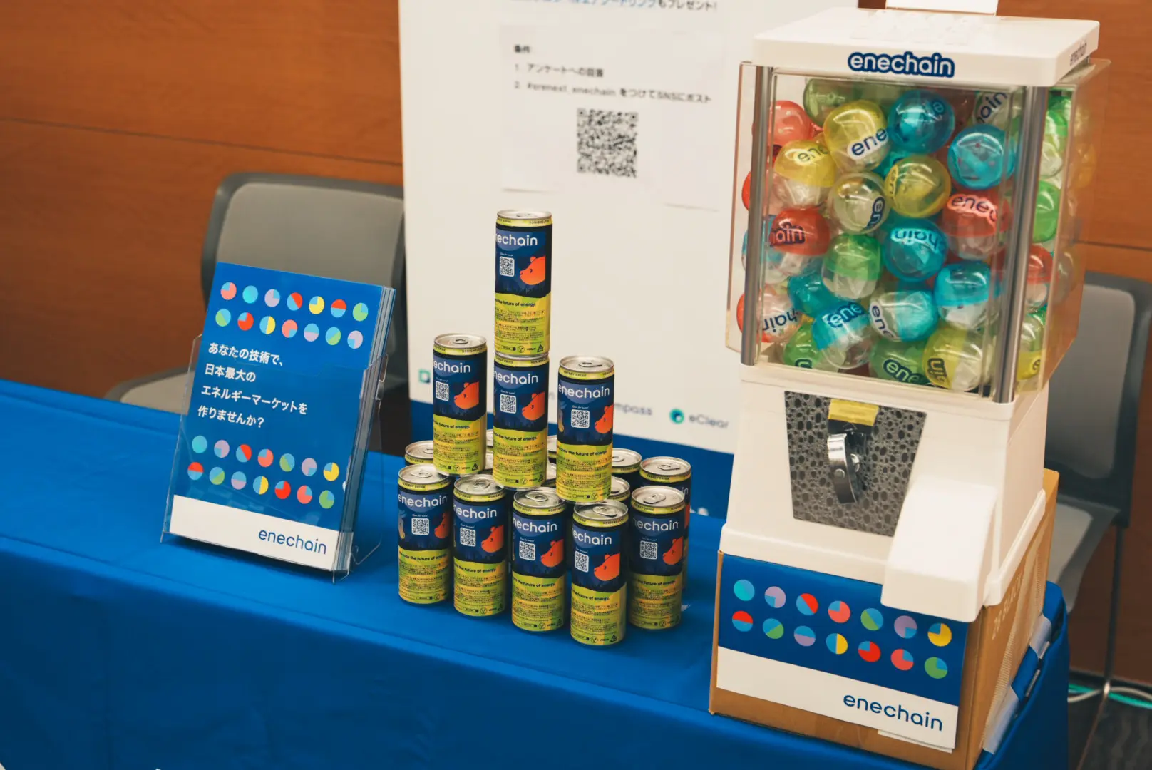 技術カンファレンスにおけるenechain社の展示ブース。青いブースクロスの上に28 BLACKオリジナルラベルエナジードリンクが積み上げられ、「日本最大のエネルギーマーケットを作りませんか？」というメッセージボードと共に展示されている